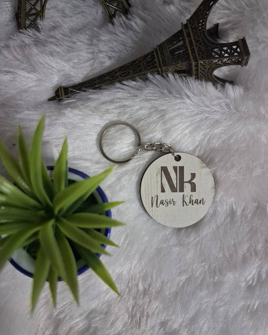 Wooden keychain with 'NK' initials on a fluffy white surface with a small plant and metal structure.